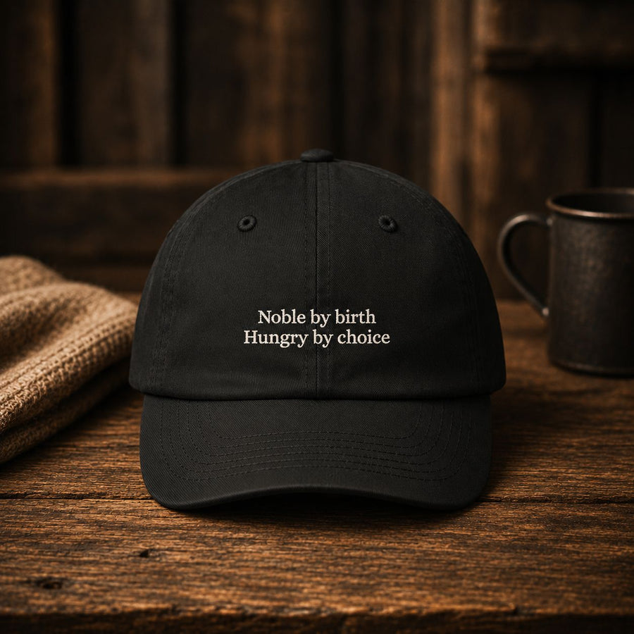 Black cotton dad cap embroidered with Noble by birth, Hungry by choice, resting on a dark wooden table beside an enamel mug